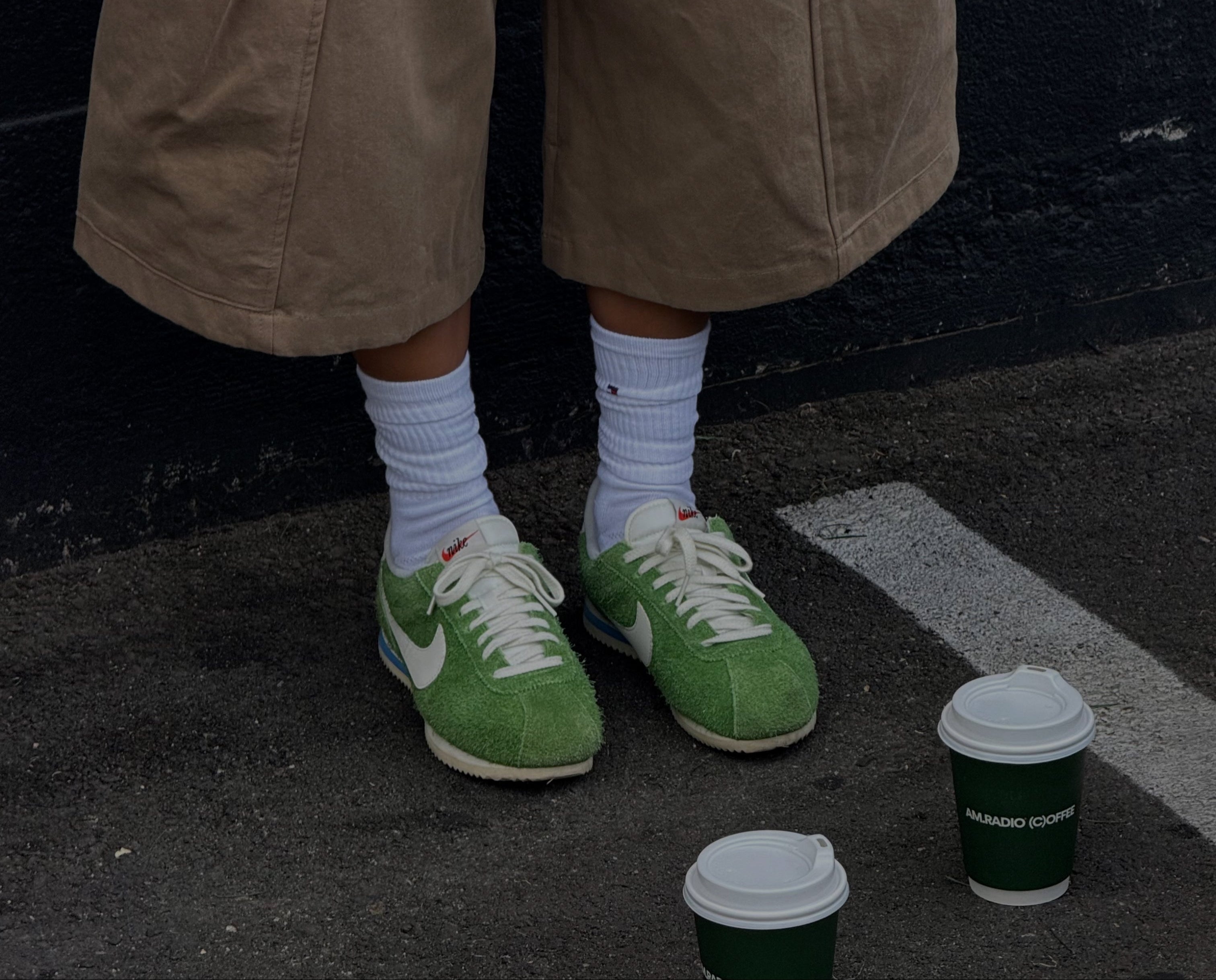 Person wearing brown cargo shorts and green sneakers with a AM.RADIO coffee cup on the ground.